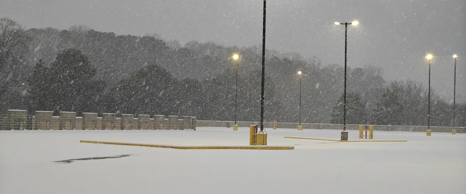 An empty parking deck under heavy falling snow at dusk — unbroken white ground, a line of lamp posts glowing sodium-yellow against grey sky and dark winter trees.